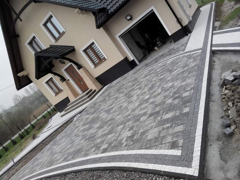 Ułożony podjazd z szarej kostki brukowej w jodełkę z białymi i ciemnoszarymi obrzeżami prowadzący do domu z garażem.