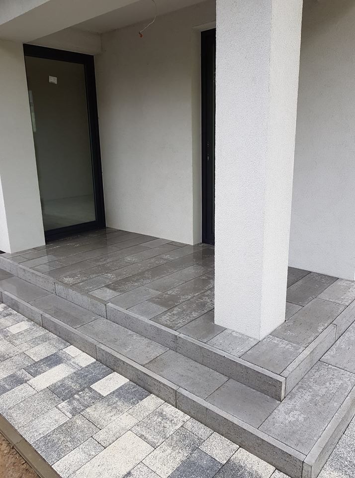 Wyłożony szarymi płytami taras z dwoma schodami, zakończony kostką w odcieniach szarości i bieli, z filarem podtrzymującym zadaszenie i czarnymi drzwiami w tle.