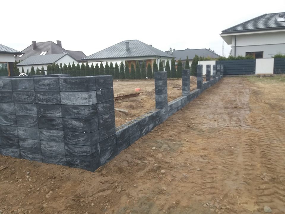 Wznoszenie muru oporowego z szarych, betonowych bloczków w surowym terenie budowy, widoczne fundamenty i przygotowany wykop pod dalszą konstrukcję, w tle domy jednorodzinne i rząd tuj.
