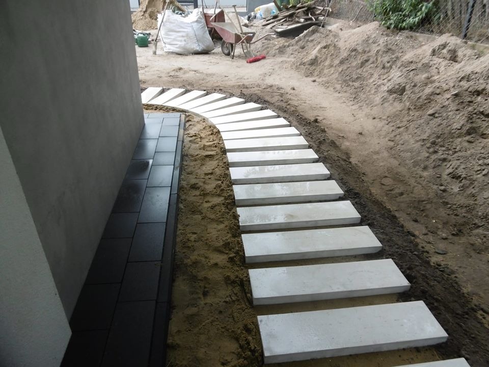 Ścieżka z betonowych płyt ułożona łukiem w piaszczystym podłożu, obok ściany budynku wyłożonej ciemnymi płytkami, w tle widoczny kopiec ziemi i narzędzia.