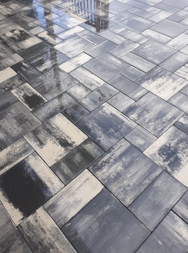 Świeżo ułożona, mokra kostka brukowa w odcieniach szarości i beżu, ułożona we wzór jodełki, odbijająca niebo i balustradę w połyskującej powierzchni.