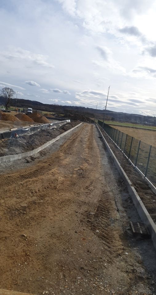 Przygotowany teren pod budowę drogi z ułożonymi obrzeżami, widoczne ślady opon, w tle hałdy piasku i maszyny budowlane, ogrodzenie z siatki po prawej stronie.