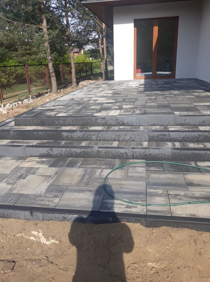 Nowo ułożony taras z szarej kostki brukowej z trzema stopniami, widoczny cień osoby, obok trawnik i ogrodzenie.