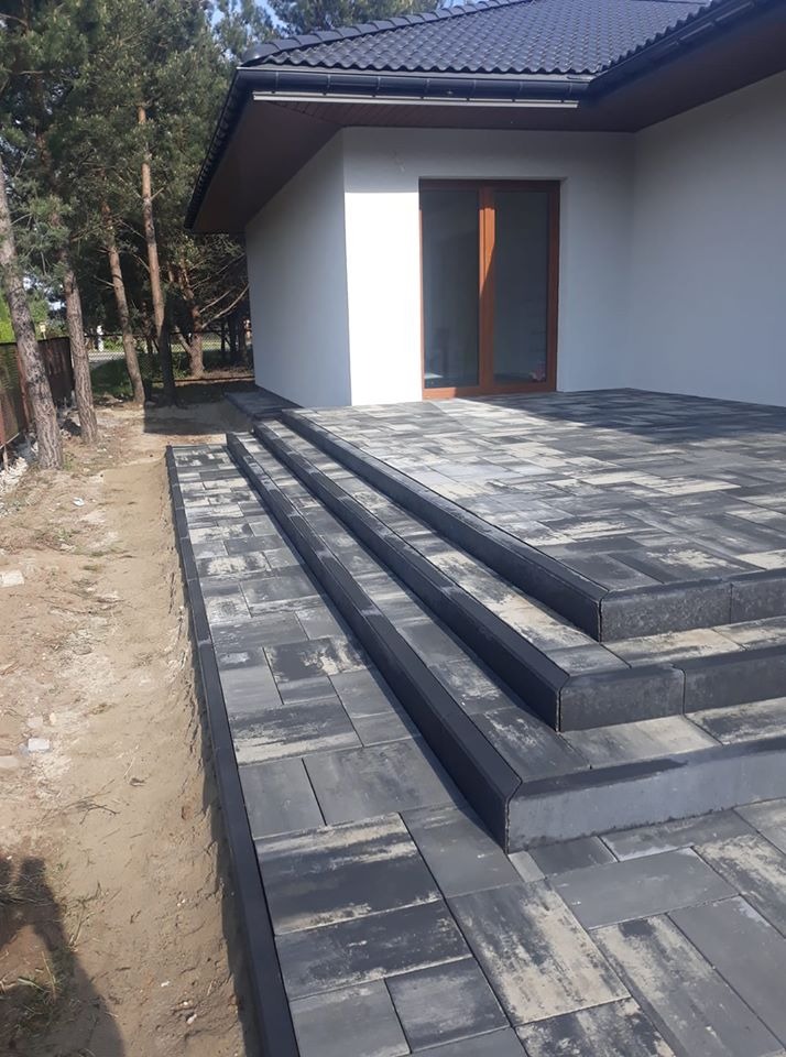 Schody i taras z szarej kostki brukowej ułożone przed nowoczesnym domem z białymi ścianami i ciemną dachówką.