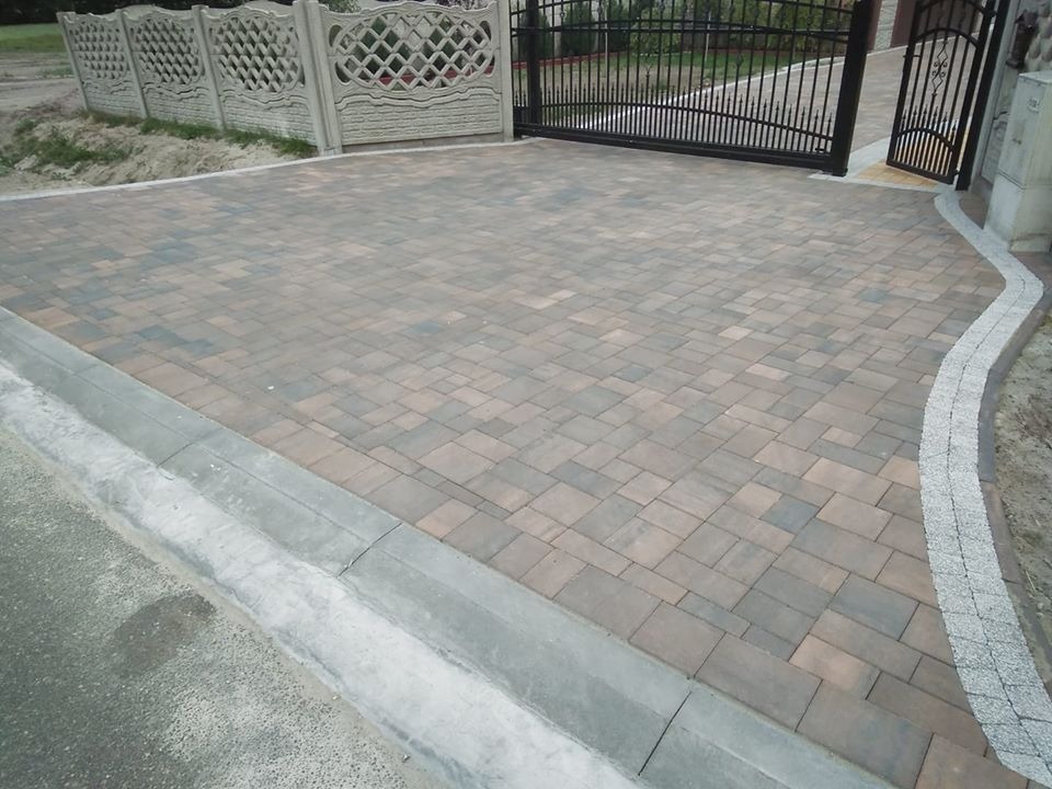 Ułożona kostka brukowa w odcieniach brązu i szarości tworząca podjazd przed domem, z widoczną żelazną bramą i betonowym ogrodzeniem.