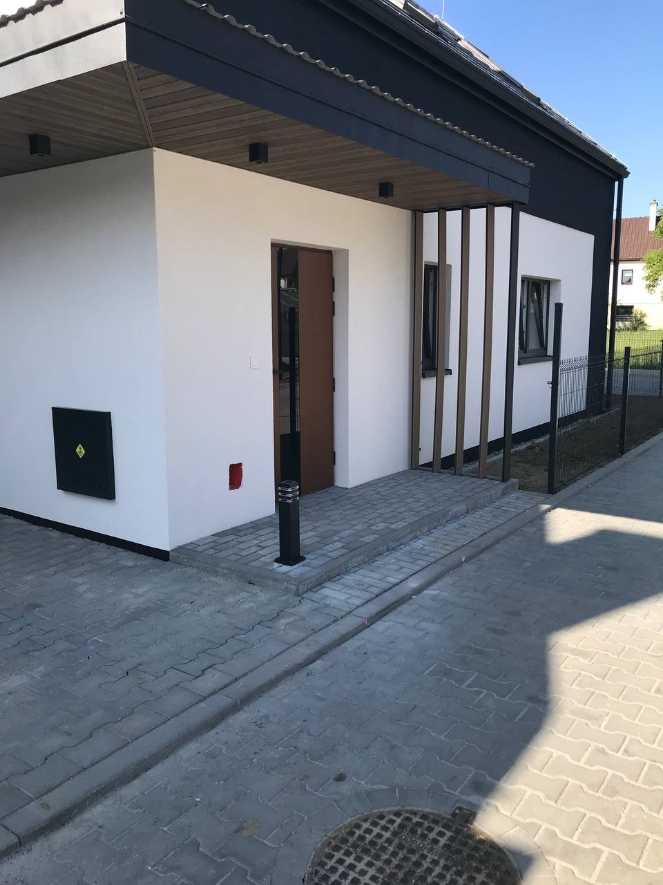 Nowoczesny budynek jednorodzinny z białą elewacją, ciemnym dachem i drewnianymi elementami dekoracyjnymi przy oknach; kostka brukowa przed wejściem.