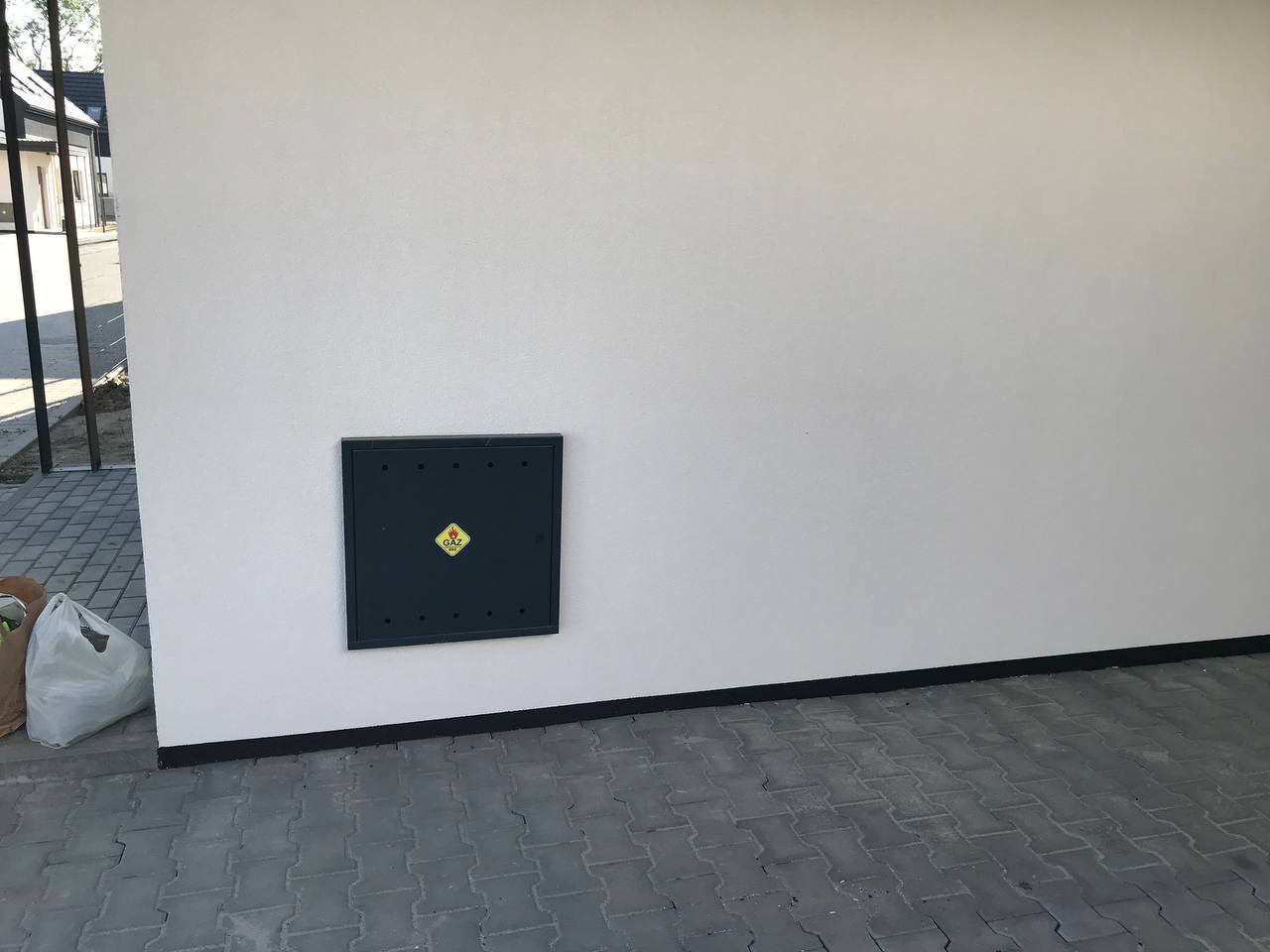 Jasna elewacja budynku z czarną skrzynką gazową i czarną listwą przypodłogową, widoczny fragment chodnika z kostki brukowej.