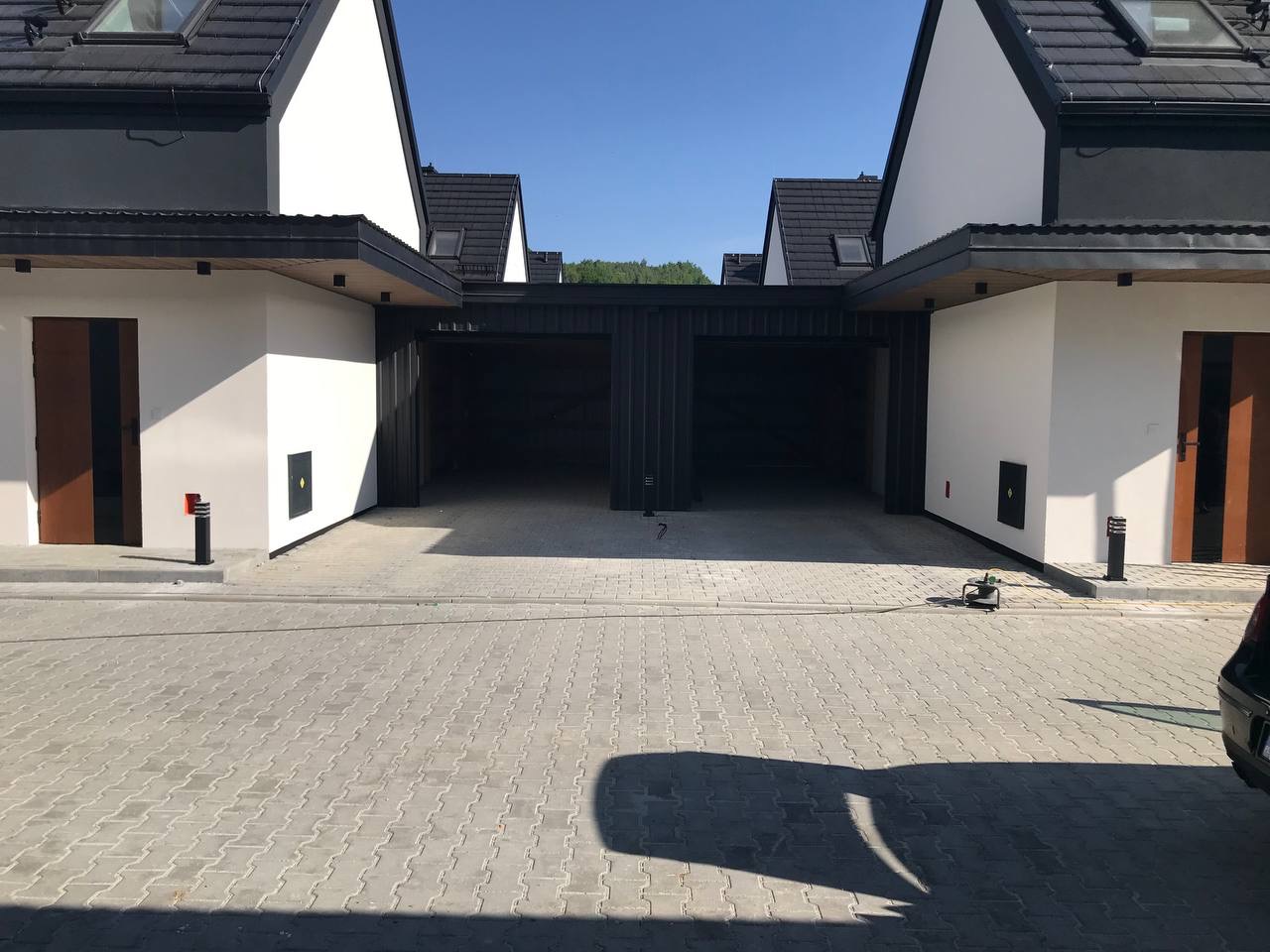 Nowoczesne szeregówki z białymi elewacjami, ciemnymi dachami i wiatami garażowymi, widok z brukowanego podjazdu, w tle zieleń drzew.