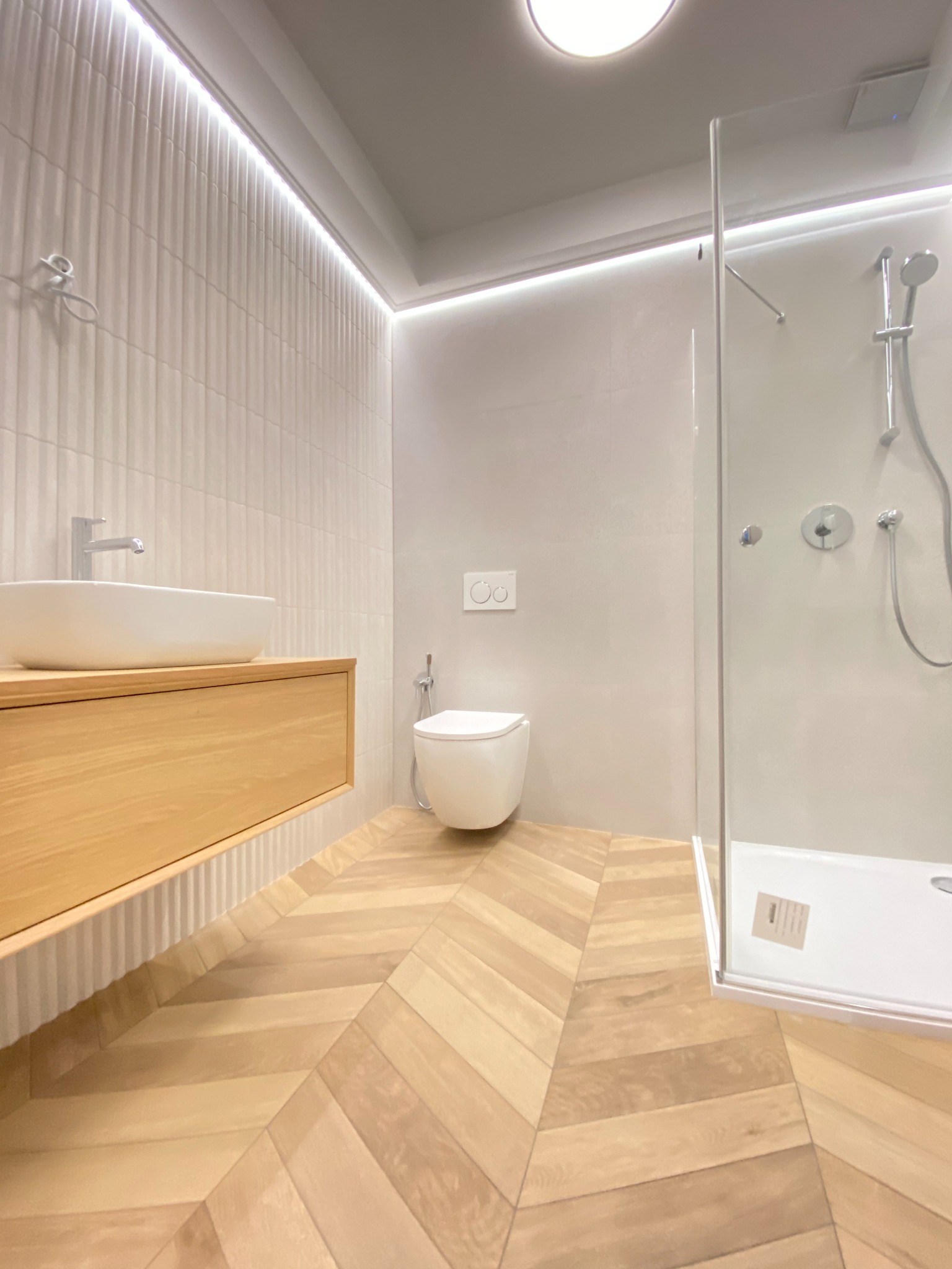 Nowoczesna łazienka z drewnianą podłogą ułożoną w jodełkę, białą umywalką nablatową na drewnianej szafce, kabiną prysznicową ze szklanymi drzwiami i toaletą podwieszaną.