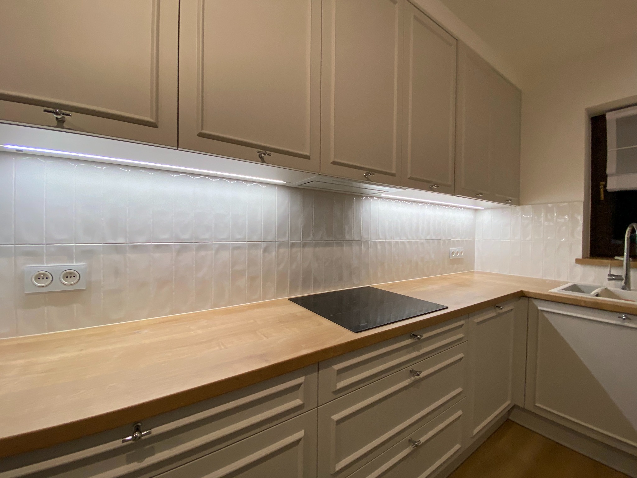 Jasna kuchnia z drewnianym blatem, płytki w odcieniach bieli, oświetlenie LED pod szafkami, płyta indukcyjna i zlewozmywak w rogu.