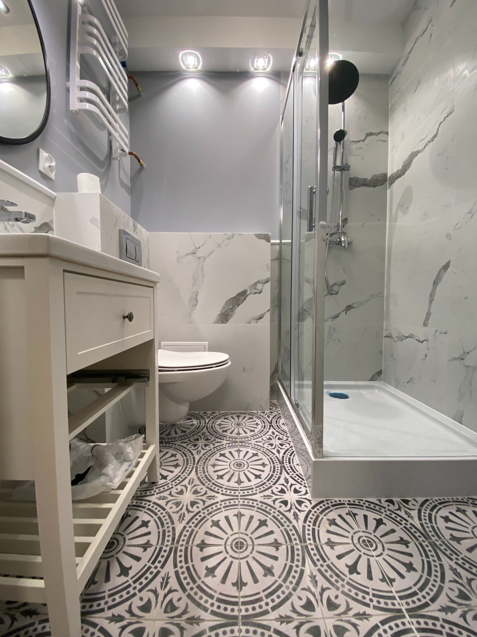 Nowoczesna łazienka z kabiną prysznicową typu walk-in, białą umywalką z szafką, toaletą i grzejnikiem drabinkowym, wykończona płytkami imitującymi marmur i wzorzystymi płytkami podłogowymi.