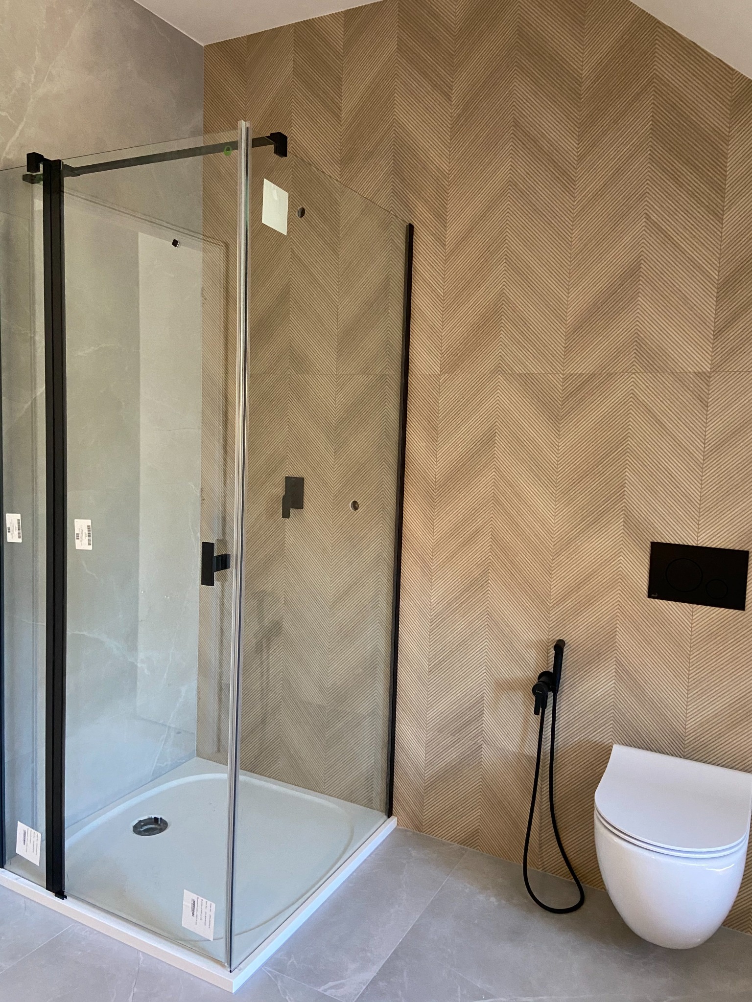 Nowoczesna łazienka z kabiną prysznicową typu walk-in z czarnymi profilami, wisząca miska WC z bidetką i geometryczną okładziną ścienną w odcieniach beżu i szarości.