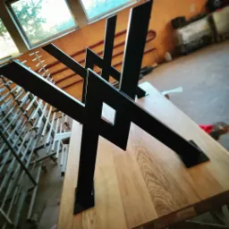 Stół z blatem z jasnego drewna i metalową, geometryczną podstawą wykonaną w Dubinach. Widoczne spawane elementy konstrukcyjne i surowe wykończenie.