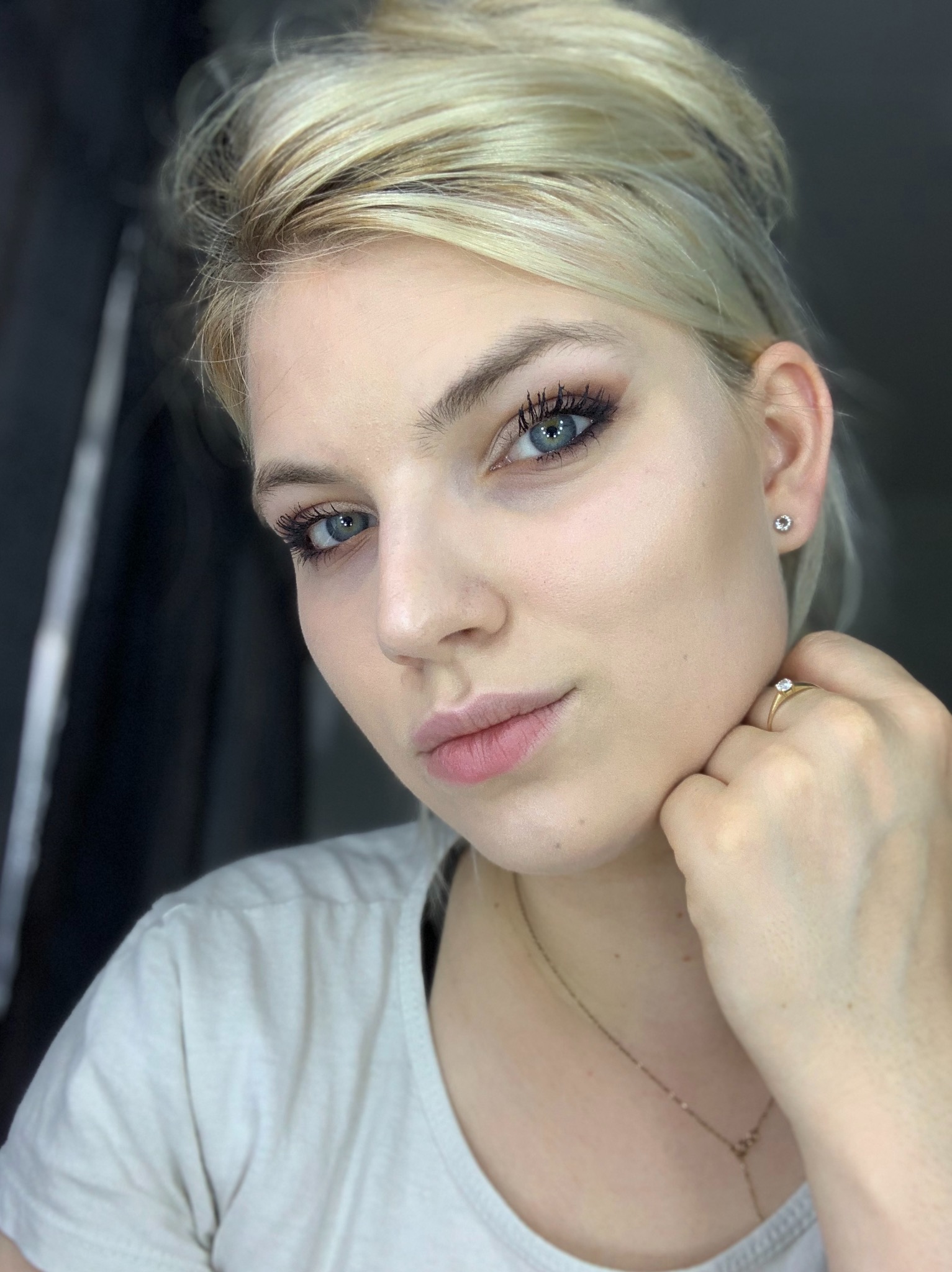Portret młodej kobiety z blond włosami upiętymi w kok, z delikatnym makijażem oczu i pierścionkiem na palcu.