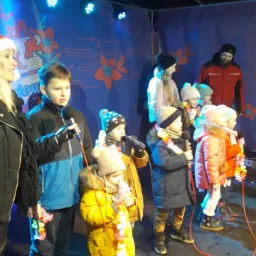 Zespół dziecięcy w zimowych ubraniach i hawajskich naszyjnikach śpiewa kolędy na scenie oświetlonej niebieskimi reflektorami, z kobietą w czapce Mikołaja i mężczyzną w tle.