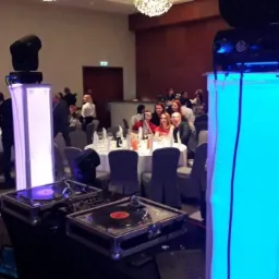 Dwa gramofony na stole DJ-skim z oświetleniem LED, kolumny głośnikowe na statywach, reflektor, projektor i bawiący się goście w tle podczas imprezy.