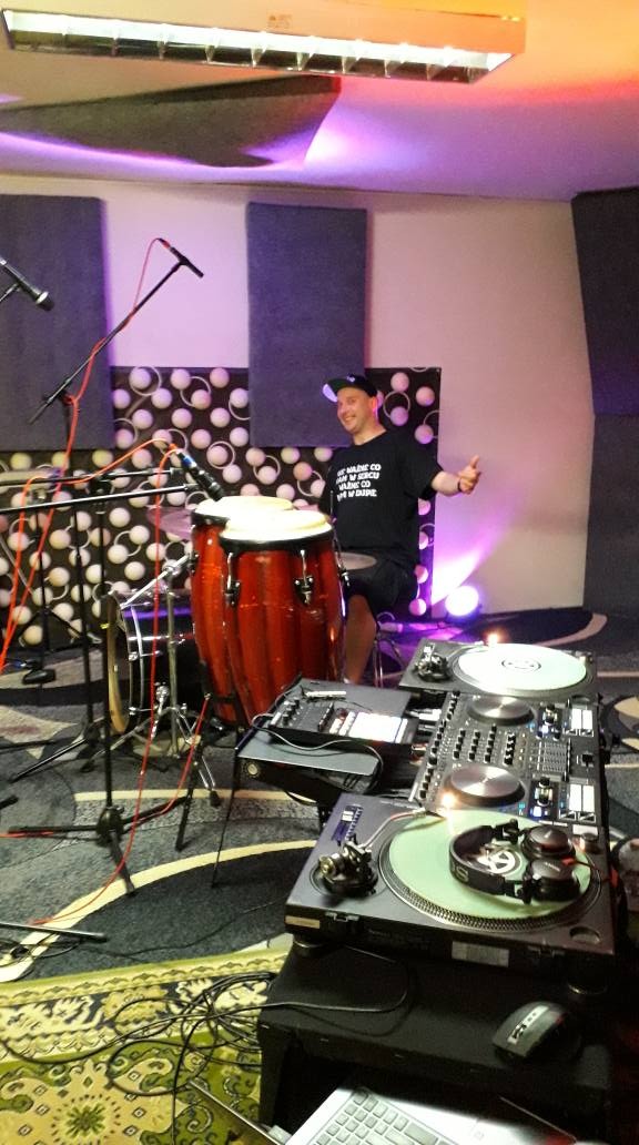 Muzyk w studiu nagraniowym, siedzący za bębnami conga i obok sprzętu DJ-skiego, uśmiechnięty i pokazujący gest aprobaty, z mikrofonami i panelami akustycznymi w tle.