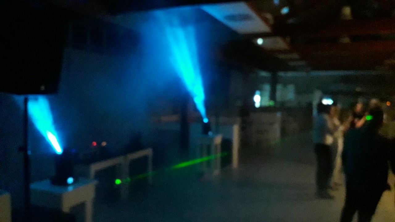 Scena z niebieskimi reflektorami punktowymi i zielonym laserem, w tle sylwetki osób, widoczny głośnik. Słaba jakość zdjęcia.
