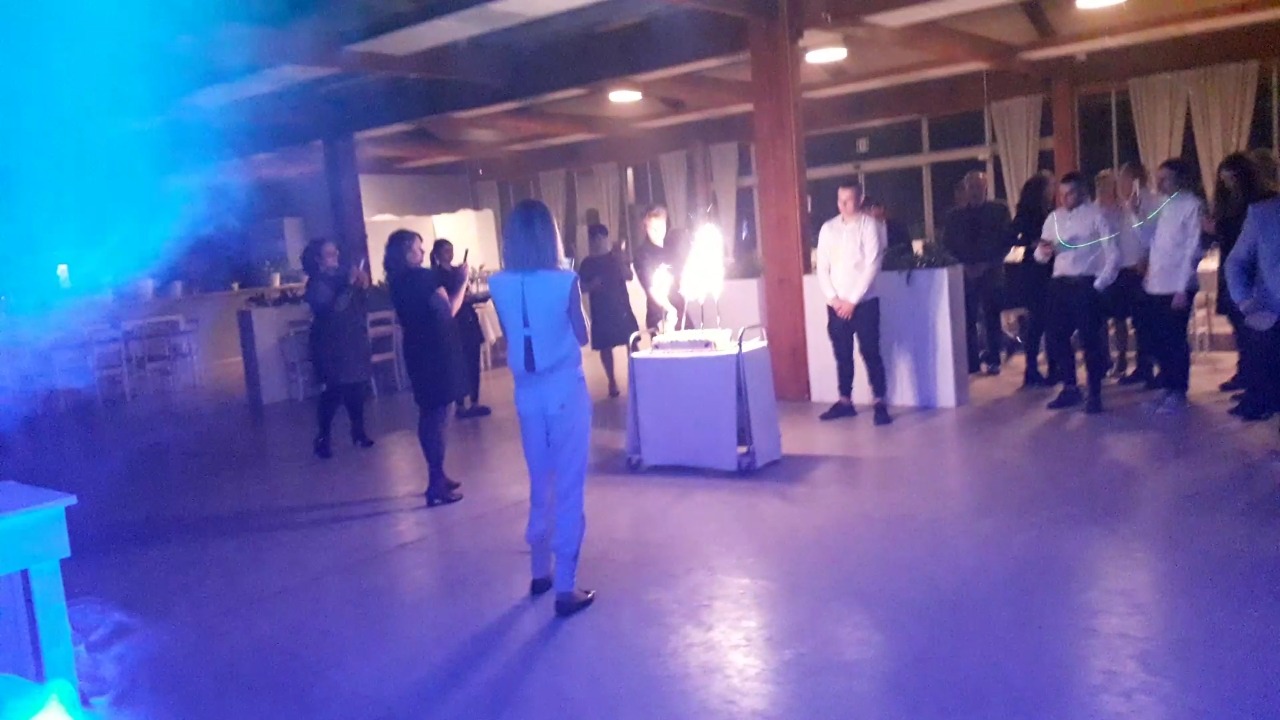 Goście weselni obserwują tort z płonącymi fontannami na wózku, oświetlony niebieskim światłem scenicznym.