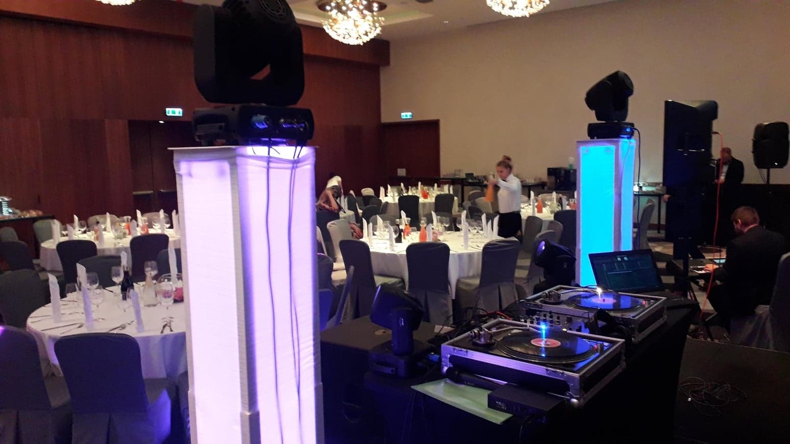 Sala bankietowa z okrągłymi stołami przygotowanymi do uroczystości, oświetlenie sceniczne na kolumnach z podświetleniem LED oraz stanowisko DJ-skie z gramofonami i laptopem.