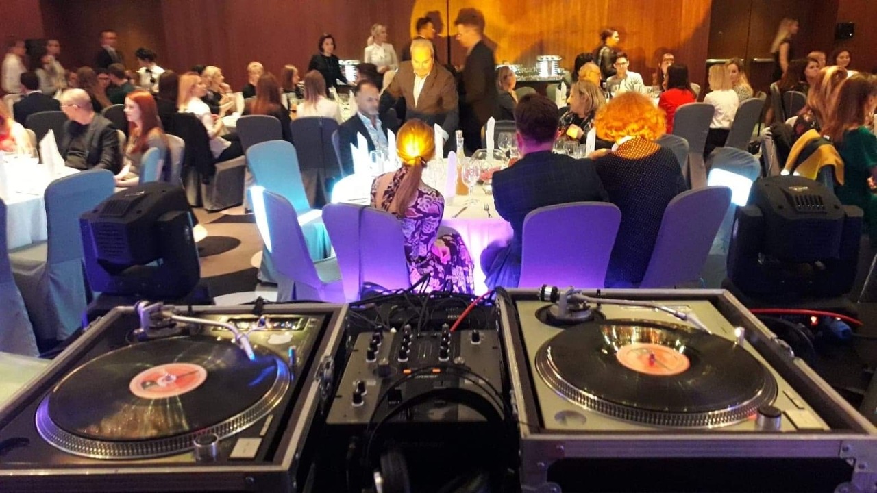 Dj stanowisko z dwoma gramofonami i mikserem na tle sali bankietowej z gośćmi siedzącymi przy stolikach, oświetlonych reflektorami.