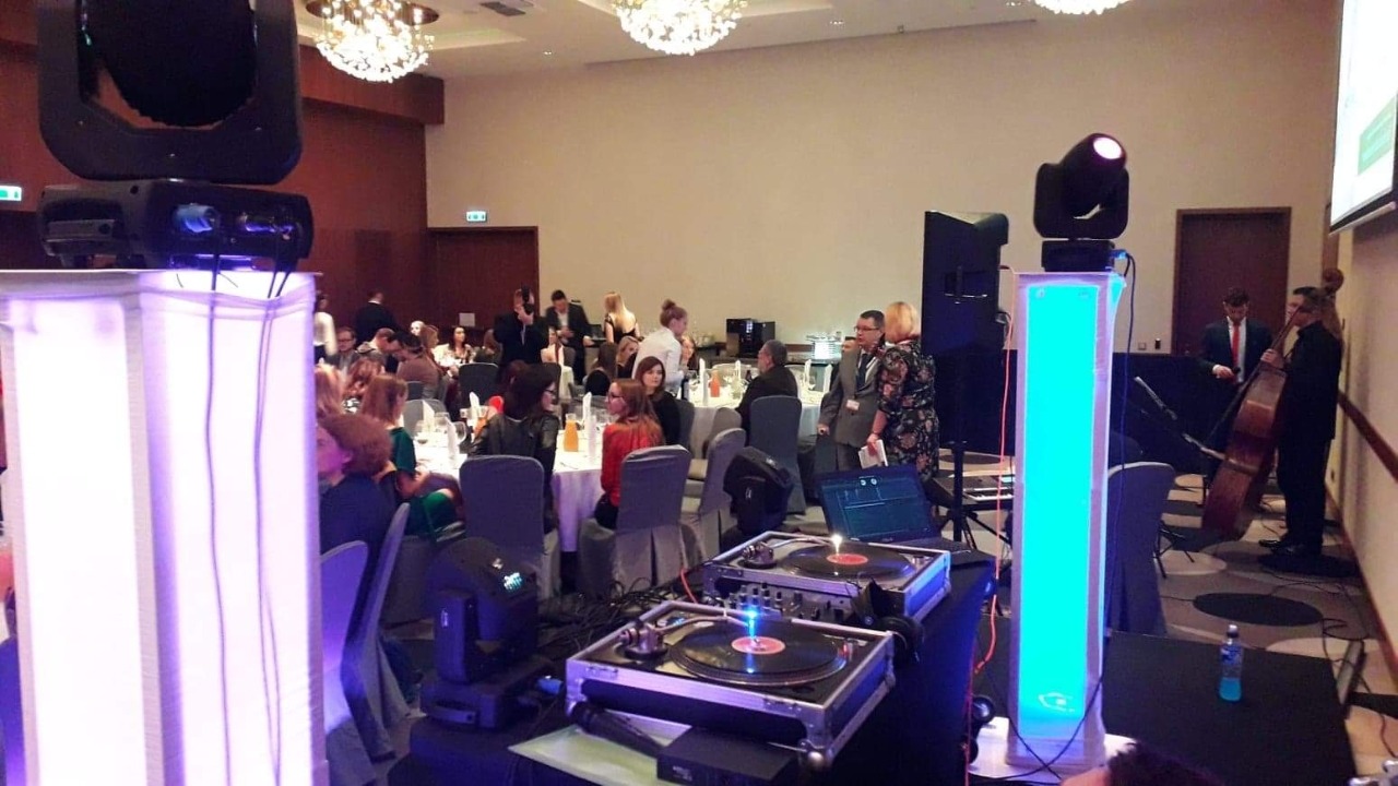 Sala bankietowa z oświetleniem scenicznym, gramofonami DJ-skimi i muzykami grającymi na kontrabasie, podczas eventu.