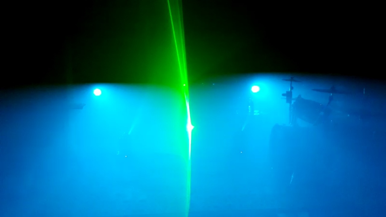 Scena oświetlona niebieskimi i zielonymi światłami laserowymi, widoczny zarys perkusji w tle.