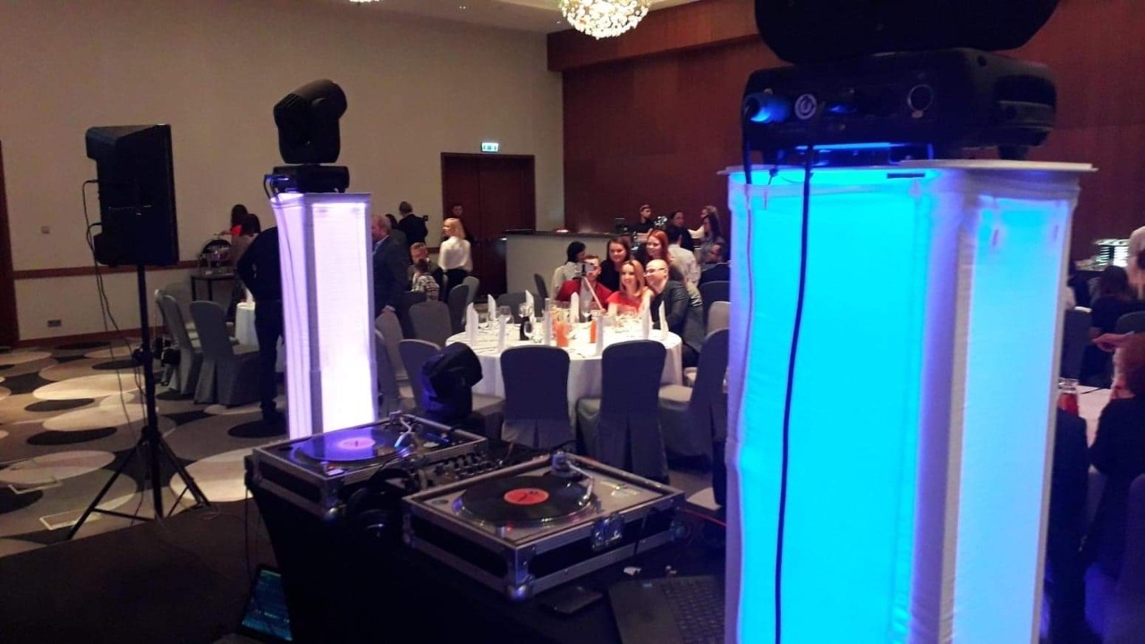 Dwa gramofony na stole DJ-skim z oświetleniem LED, kolumny głośnikowe na statywach, reflektor, projektor i bawiący się goście w tle podczas imprezy.