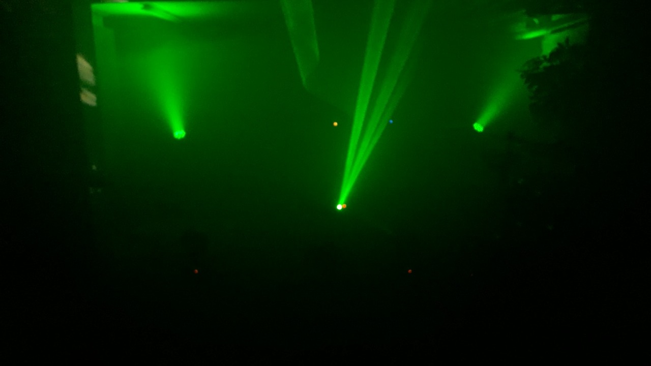 Scena oświetlona zielonymi reflektorami, z widocznymi promieniami światła w ciemności, tworzącymi atmosferę imprezy.
