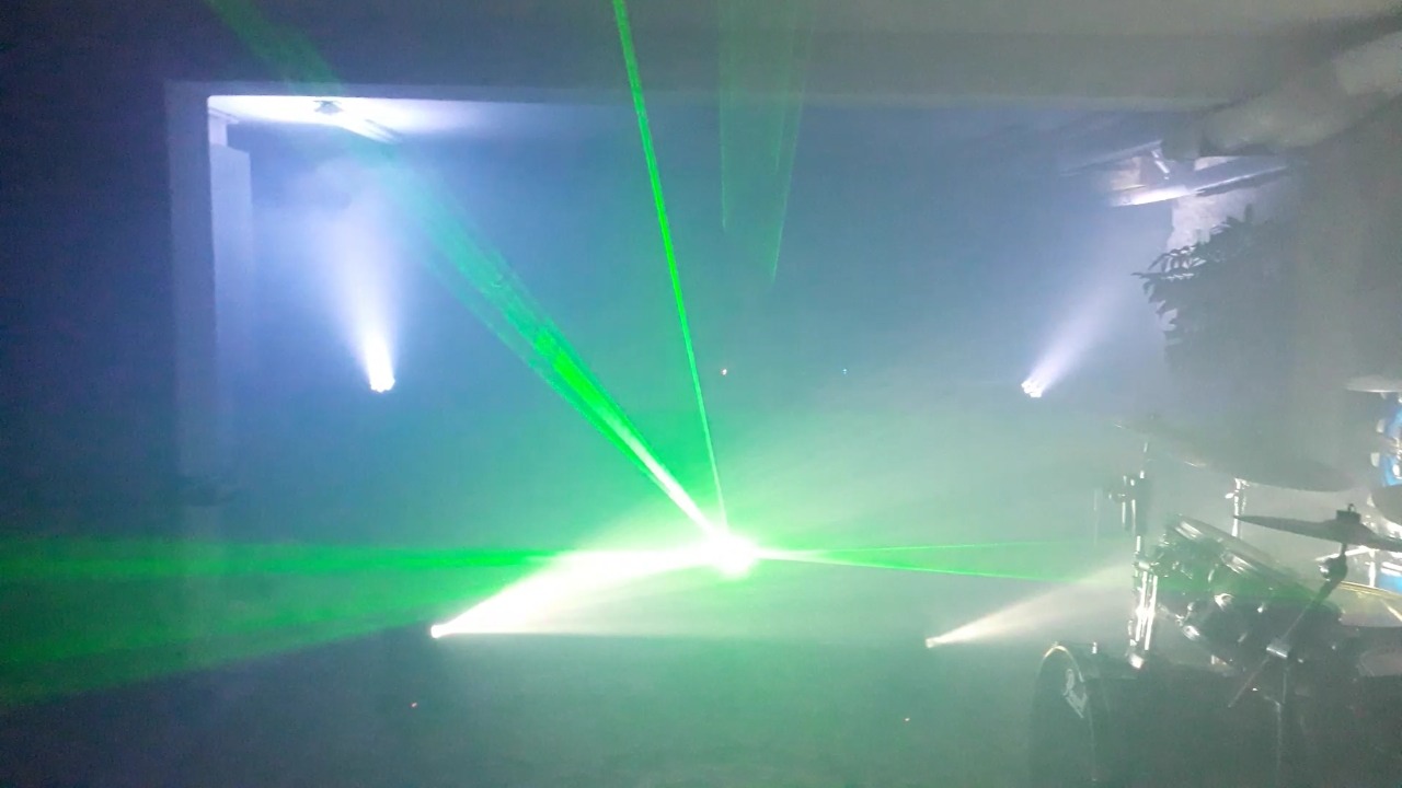 Scena z efektami świetlnymi: zielone lasery przecinają dym, oświetlając perkusję w tle.