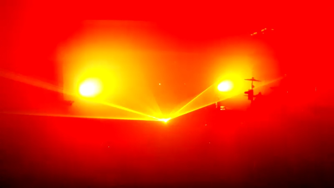 Scena oświetlona intensywnym czerwonym światłem, z widocznymi reflektorami i zarysem perkusji w tle.