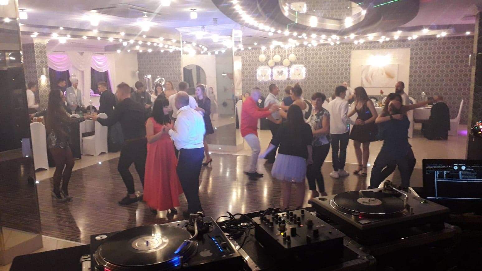 Parkiet taneczny podczas imprezy, widoczny sprzęt DJ-ski: gramofony i mikser, w tle tańczący goście w różnym wieku i strojach.