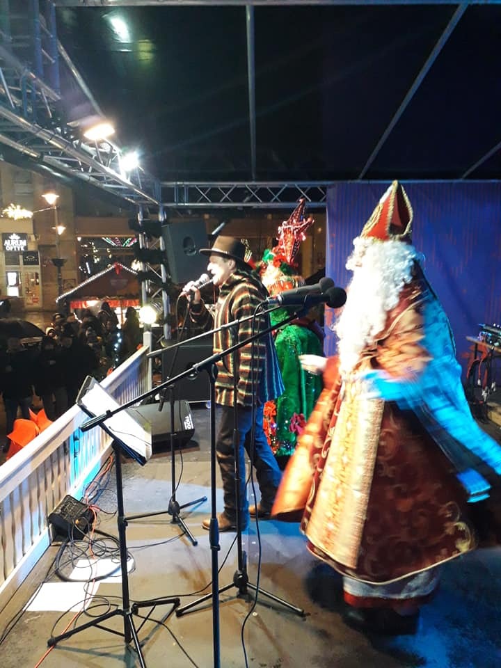 Występ na scenie: wokalista w kapeluszu śpiewa do mikrofonu, obok postacie w kostiumach świątecznych, publiczność w tle.