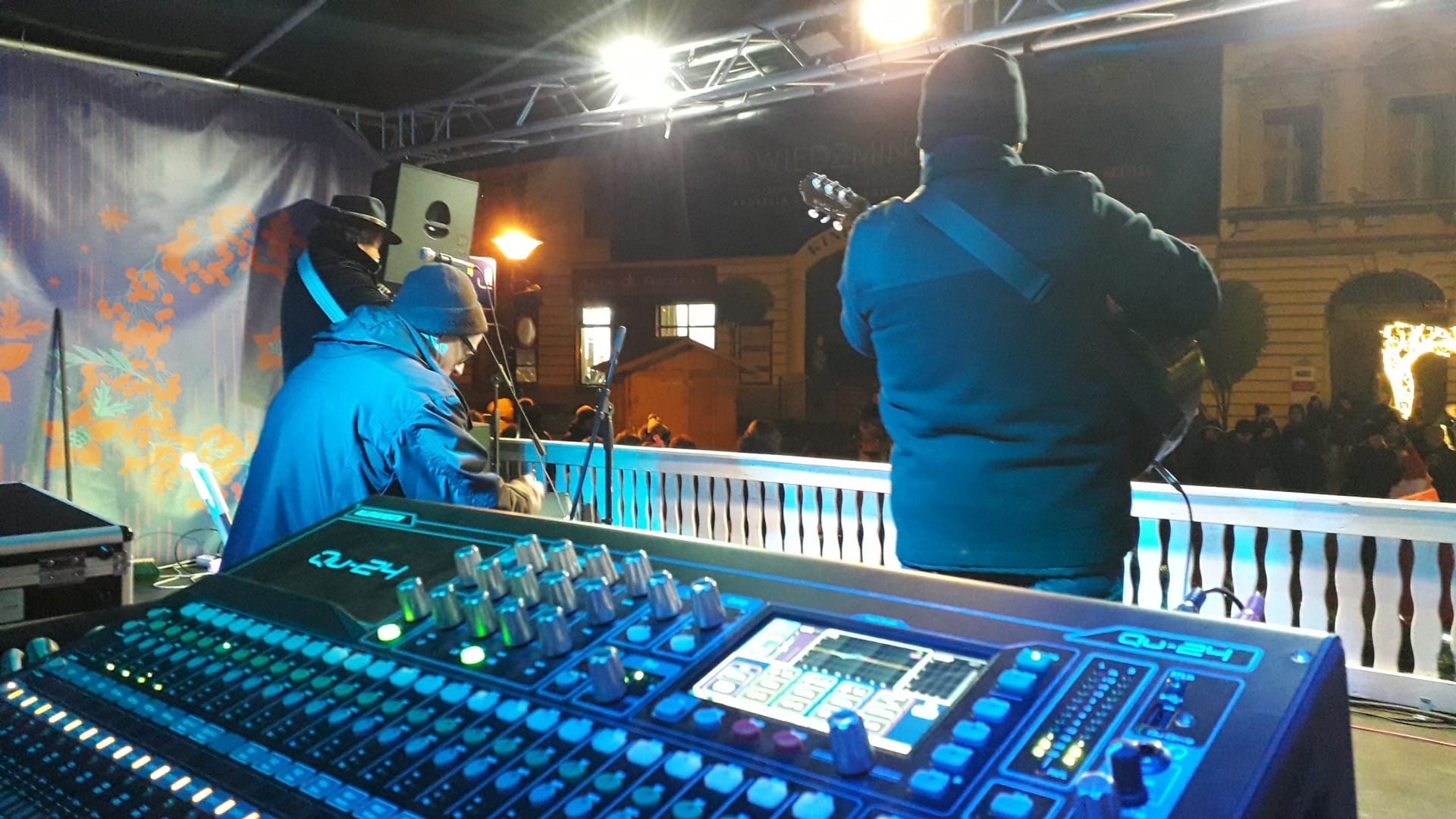 Koncert plenerowy nocą: widok na mikser dźwięku Qu-24, muzyków na scenie i publiczność w tle, oświetlenie sceniczne.