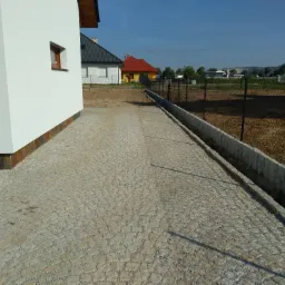 Ułożona kostka granitowa tworząca podjazd obok białego domu z brązowymi elementami wykończeniowymi, widoczne ogrodzenie i przygotowany teren pod trawnik.