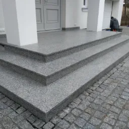 Trzystopniowe schody wejściowe z szarego granitu, prowadzące do drzwi wejściowych, z kostką brukową na podłożu i pracownikiem w tle.