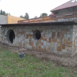 Kamienna konstrukcja z dwoma okrągłymi otworami, prawdopodobnie piwnica lub schron, wykonana z nieregularnych, szaro-brązowych kamieni, z drewnianym zadaszeniem i elementami drewnianej konstrukcji...