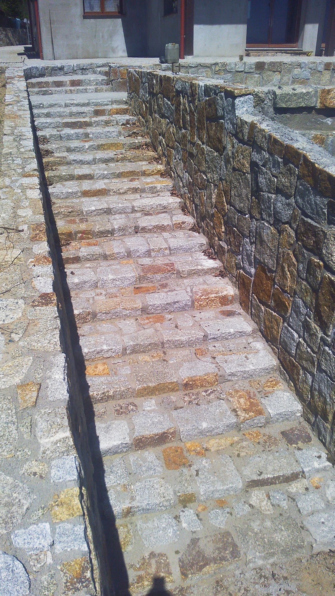 Zewnętrzne schody i mur oporowy wykonane z kostki o nieregularnych kształtach w odcieniach szarości i brązu, prowadzące do domu. Widoczny cień osoby robiącej zdjęcie.