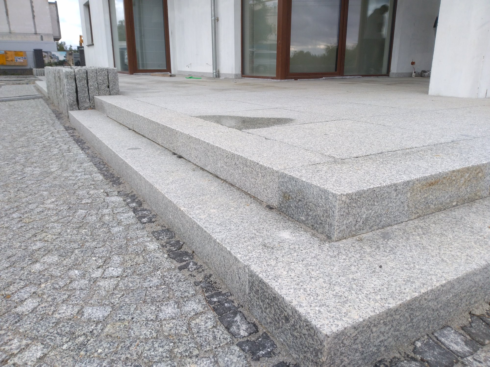 Dwu-stopniowe schody kamienne z jasnego granitu, prowadzące do budynku z dużymi oknami, obok ułożona kostka granitowa z ciemną obwódką.