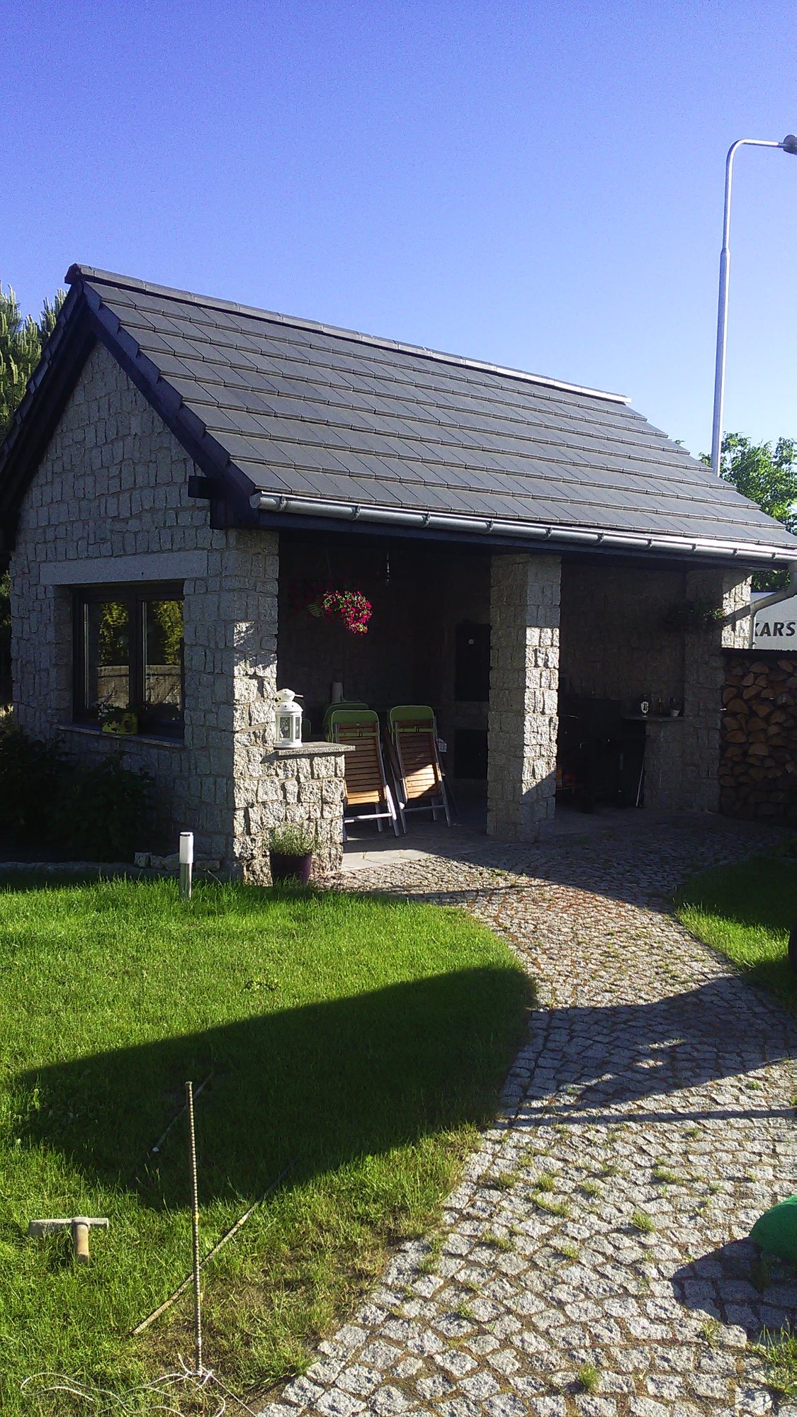 Altana z kamienia z szarą dachówką, otoczona trawnikiem i ścieżką z drobnej kostki, w tle widoczne drzewa i słup oświetleniowy.