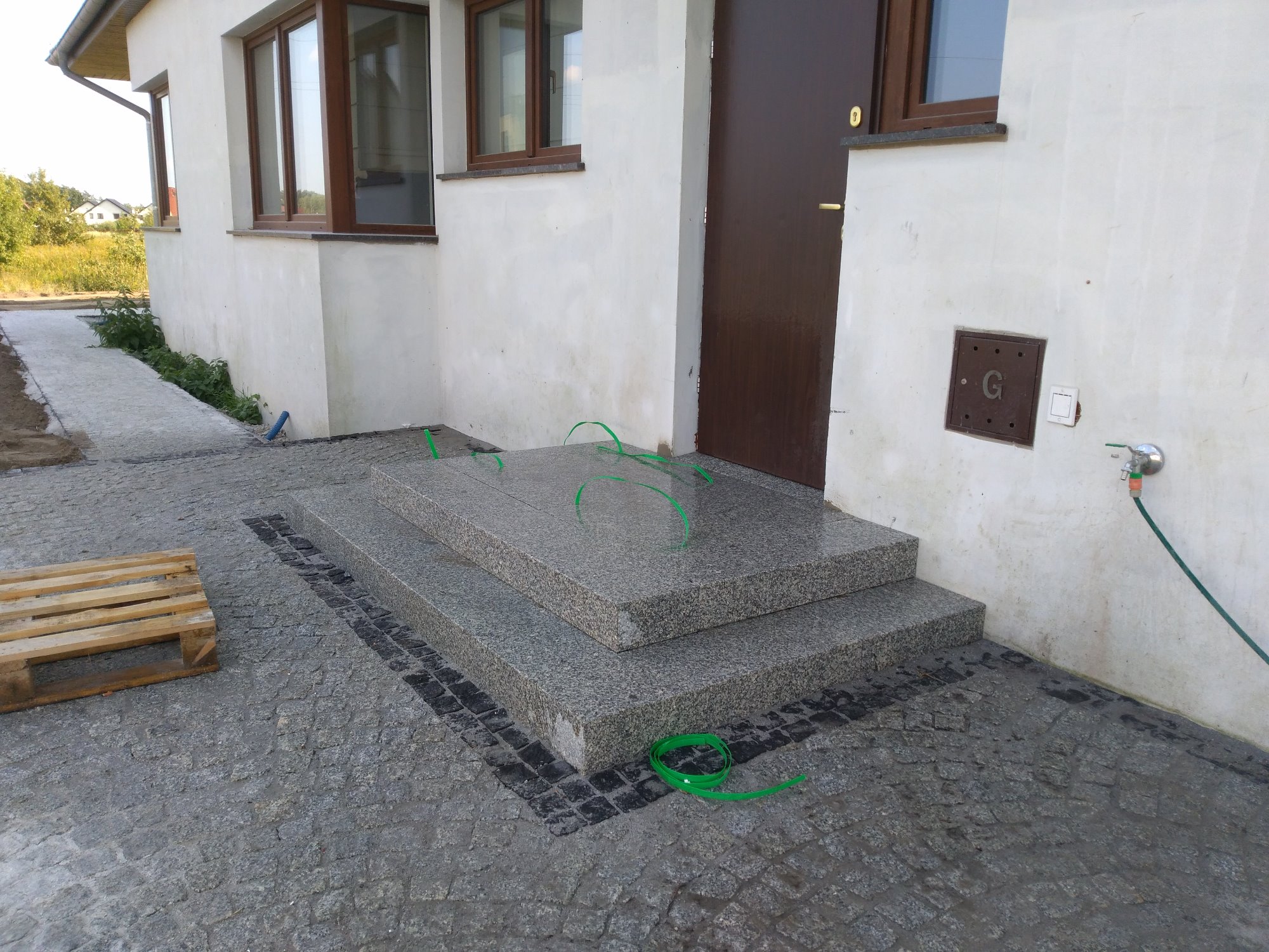 Nowe, dwustopniowe schody wejściowe z szarego kamienia, otoczone kostką brukową w odcieniach szarości, z paletą drewnianą w tle i fragmentem elewacji budynku.