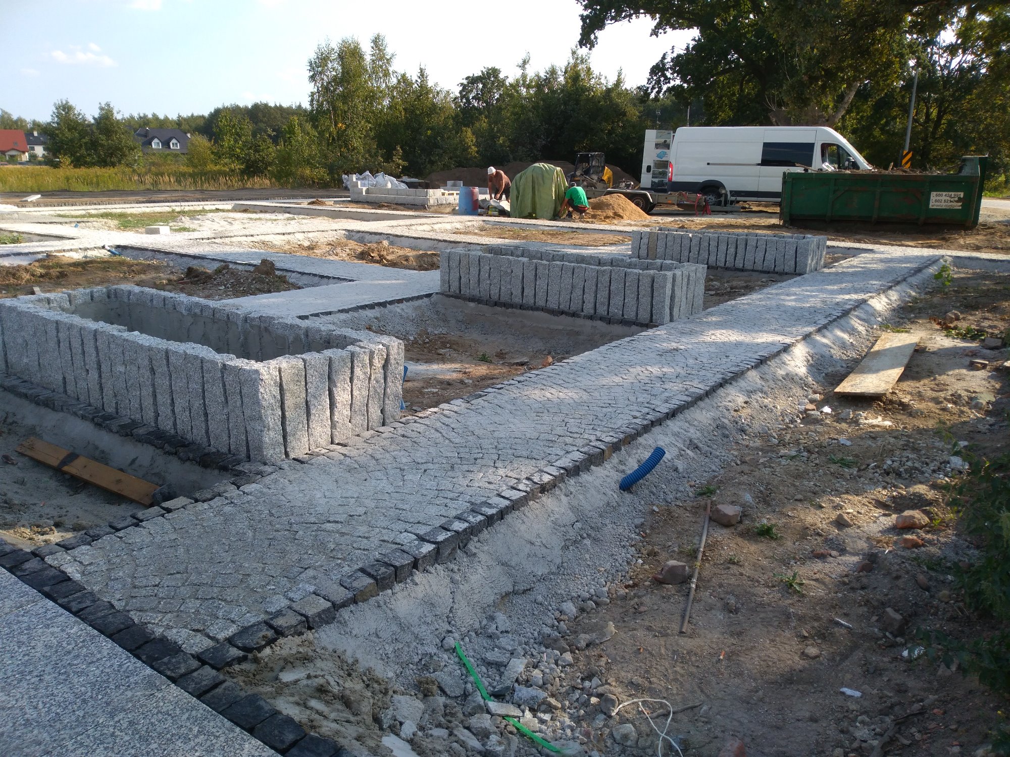 Fragment placu budowy z częściowo ułożoną nawierzchnią z kostki brukowej, widoczne fundamenty pod donice z kamienia, w tle pracownicy i kontener na gruz, częściowo przygotowany teren pod dalsze prace.