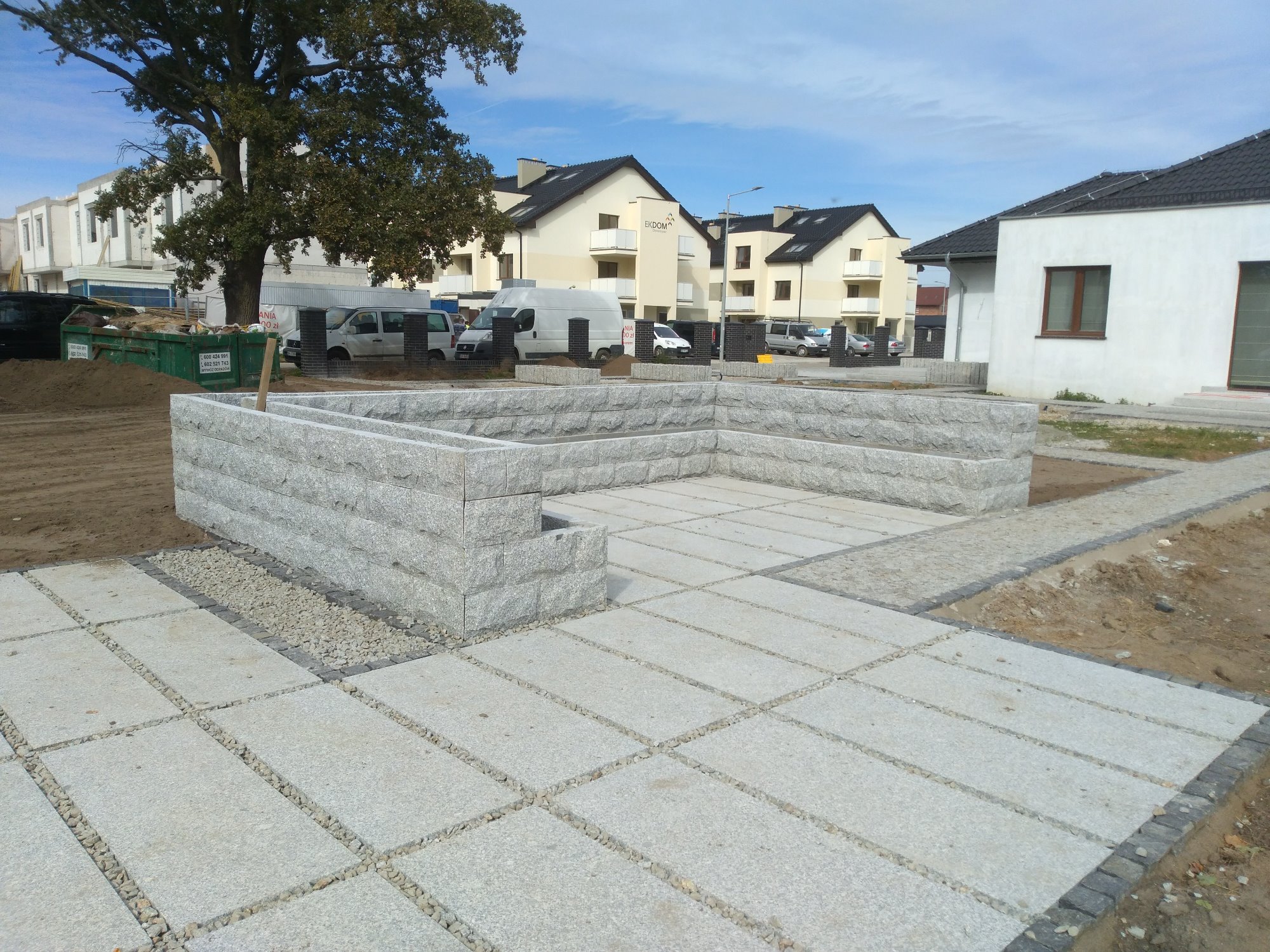 Nowo wybudowany taras z szarej kostki granitowej i kamiennych murków w nowoczesnym osiedlu, z widocznymi nowymi domami w tle, jeszcze w trakcie prac wykończeniowych.