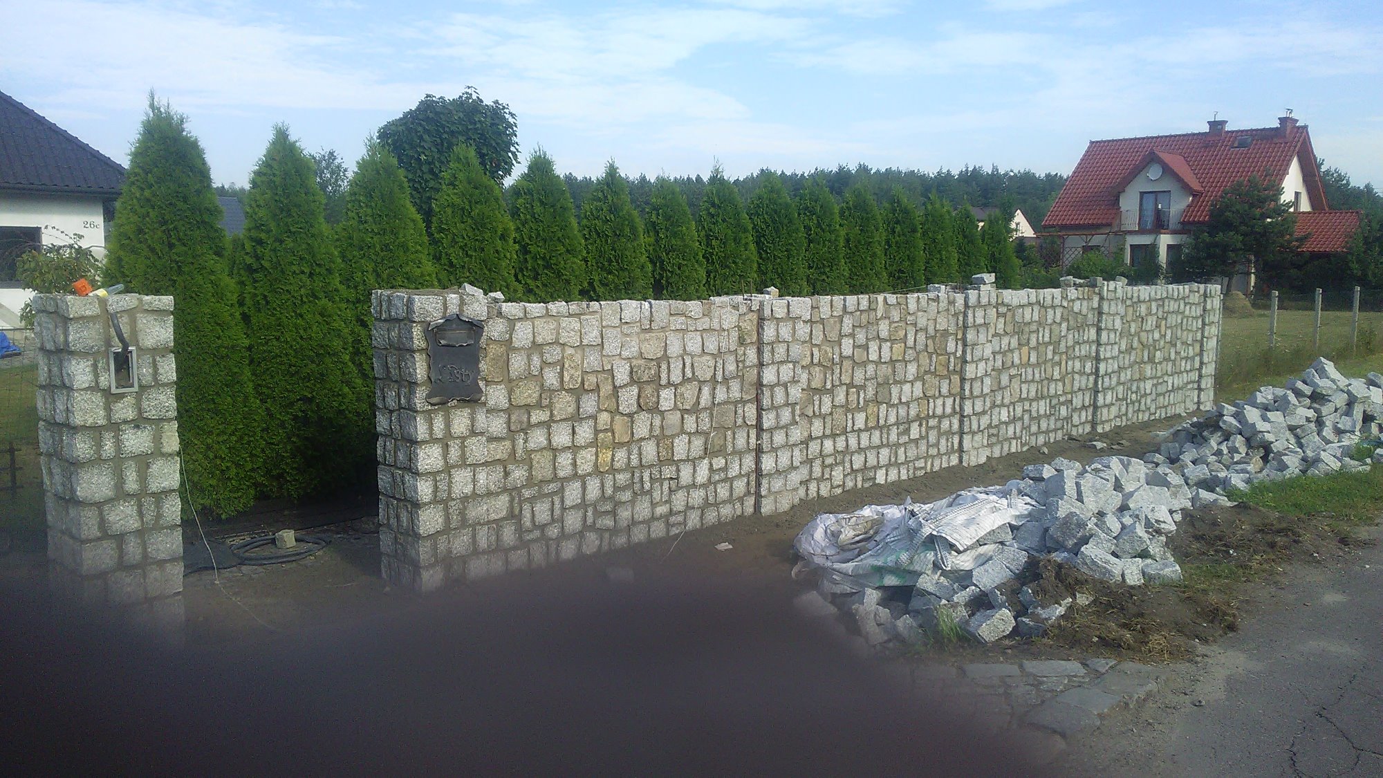 Wznoszenie ogrodzenia z nieregularnej kostki granitowej w odcieniach szarości, z widocznymi słupkami wejściowymi i stertą nieużytych kamieni obok, na tle rzędu wysokich tuj i domu z czerwoną dachówką.