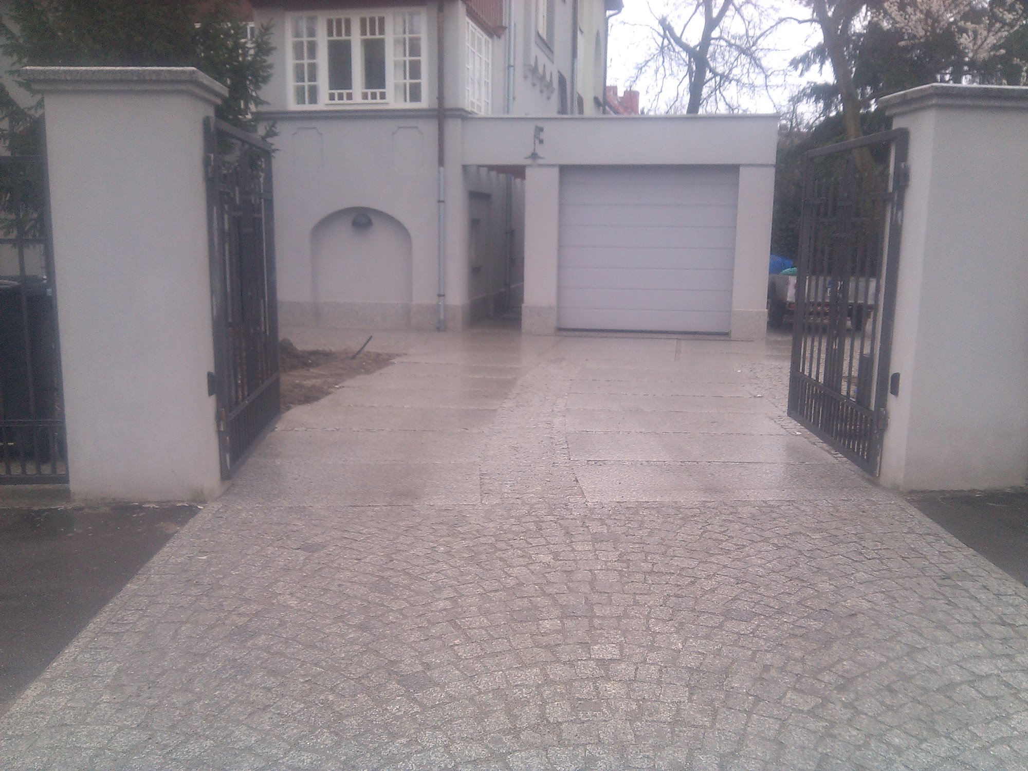 Podjazd do garażu wyłożony kostką granitową w odcieniach szarości, z otwartą, kutą bramą wjazdową i budynkiem w tle, po deszczu.