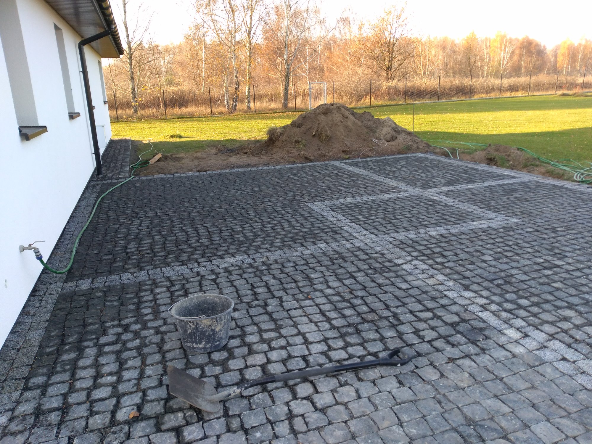 Ułożona nawierzchnia z kostki granitowej w szarych odcieniach, obok biały budynek, widoczne wiadro, łopata i pryzma ziemi na trawniku w jesiennej scenerii.