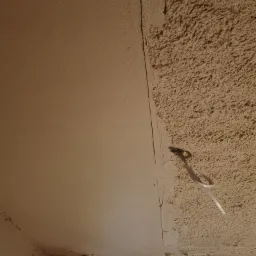 Fragment sufitu z widoczną różnicą w fakturze: gładka powierzchnia po lewej stronie i chropowata, nierówna struktura po prawej, z wystającym przewodem elektrycznym.