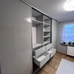 Szara szafa wnękowa z przesuwnymi drzwiami, otwarte szuflady i półki, widok na okno z firanką i grzejnik