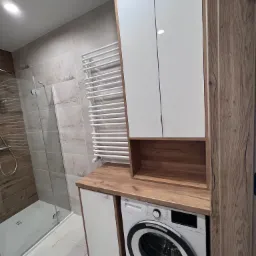 Nowoczesna łazienka z zabudową na wymiar: białe szafki z drewnianymi akcentami, pralka pod blatem, grzejnik drabinkowy i kabina prysznicowa.