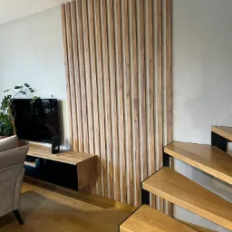 Drewniana ścianka działowa z pionowych listew w salonie z drewnianymi schodami i podłogą, obok szafka RTV i roślina doniczkowa.